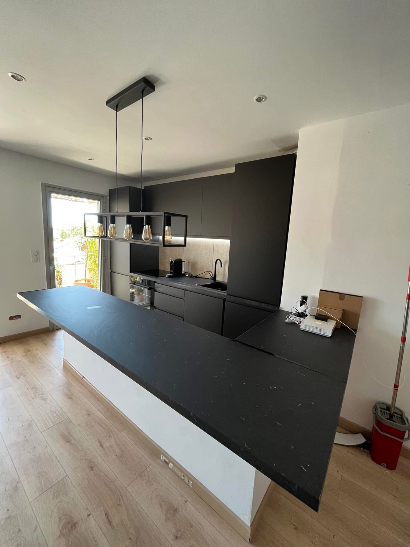 LUX RENOV — cuisine noire avec crédence pierre, plan granit et suspension industrielle